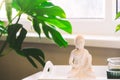 Decorative white Buddha statuette figure with candles standing on the tray near the window with a green monstera plant Royalty Free Stock Photo