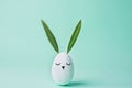 Decorative White Painted Easter Egg Bunny with Drawn Cute Kawaii Face. Green Leaves as Ears. Pastel Turquoise Background. Royalty Free Stock Photo