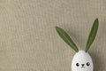 Decorative White Painted Easter Egg Bunny with Drawn Cute Kawaii Smiling Face. Green Leaves Ears. Beige Linen Fabric Background Royalty Free Stock Photo