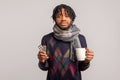 Deeply frustrated african guy with dreadlocks in checkered scarf holding cup of tea and pills in hands sadly looking at camera,