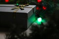 Defocus gray gift with gold bow ribbon on pine or fir tree blurred background with glowing festive green, red light bulb Royalty Free Stock Photo