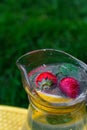 Defocus two strawberry, slice lemon and leaves of mint in glass jug of lemonade on yellow board. Blurred grass