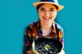 Defocus young woman in a hat and shirt smiles and shows a basket of blueberry and bilberry. Light blue background