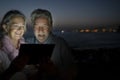 Defocused elderly woman and man sitting on the beach with laptop computer illuminating their faces. Dark night and illuminated sea Royalty Free Stock Photo