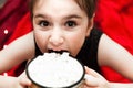 Defocused Funny little Girl Holding Cup with Hot Tasty Chocolate and Marshmellow and trying to eat it on cozy Red