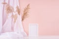 Delicate home decor with blank wooden poster frame, silk curtain and fluffy reeds in vase on white wooden table and pastel pink. Royalty Free Stock Photo