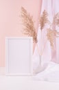 Delicate home decor with blank wooden poster frame, silk curtain and fluffy reeds in vase on white wooden table, pastel pink wall. Royalty Free Stock Photo