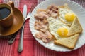 Delicious Breakfast - a Cup of coffee, a plate of fried eggs, bacon and toast, next to the Cutlery on red checkered napkin Royalty Free Stock Photo