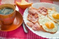 Delicious Breakfast - a Cup of coffee, a plate of fried eggs, bacon and toast, next to the Cutlery on red checkered napkin Royalty Free Stock Photo