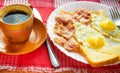 Delicious Breakfast - a Cup of coffee, a plate of fried eggs, bacon and toast, next to the Cutlery on red checkered napkin Royalty Free Stock Photo