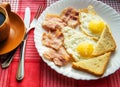 Delicious Breakfast - a Cup of coffee, a plate of fried eggs, bacon and toast, next to the Cutlery on red checkered napkin Royalty Free Stock Photo