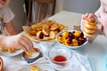 Delicious breakfast with tiny mini pancakes, cherries, apricots, jam on a white table. Small children hold pancakes in