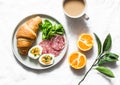 Delicious brunch or breakfast - croissant, egg, salami sausage, green salad, orange and coffee with milk on a light