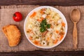 Delicious chicken soup with vegetables and noodles on a textured burlap with slices of toast, a wooden spoon and cherry tomatoe