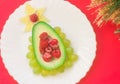 Delicious edible Christmas tree of avocado, frozen sweet raspberries and grapes on a white plate on a red table near the branches Royalty Free Stock Photo