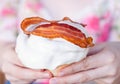 Delicious freshly baked cinnamon roll covered in white frosting with a piece of bacon on top held by hands in close up Royalty Free Stock Photo