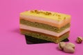 Delicious multi-layer pastry sitting next to a couple of pistachios on a pink background