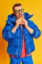 Delicious. Portrait of handsome man in blue winter jacket posing with burger over bright yellow background. Concept of