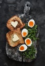 Delicious simple breakfast - boiled eggs with soft yolk, arugula, toast with butter on a rustic cutting board on a dark background Royalty Free Stock Photo