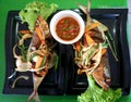 The deliciousness of two fried fish in a black plate, served with Budu water and various vegetables on a green background.