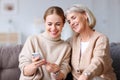 Delighted adult and aged women smiling and browsing social media on smartphone while resting on couch at home