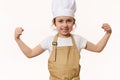 Overjoyed baby girl dressed as confectioner in chef's hat and apron clenches fists, looks at camera, on white Royalty Free Stock Photo