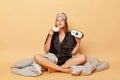 Delighted satisfied young girl sitting floor with blanket wearing black pajama and sleep eye mask holding alarm clock and cup of Royalty Free Stock Photo