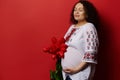 Delightful adult pregnant woman in Ukrainian ethnic clothing, holding belly, posing with red tulips, background