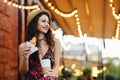 Delightful brunette female with pleasant appearance wearing summer hat and dress holding croissant and takeaway coffee, resting at