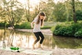 Delightful little female athlete happily jumping on green mat on sandy beach near river. Girl doing sports outdoors. Royalty Free Stock Photo