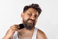 Delightful mature man taking care after beard, shaving with shaver against white studio background. Home spa