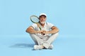 Delightful. Portrait of handsome senior man in stylish white outfit sitting, posing with tennis racket over blue Royalty Free Stock Photo