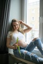 Delightful preggers woman with bare tummy sitting on sill with tulip in hand, looking at flower and smiling happily. Royalty Free Stock Photo