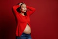 Delightful pregnant woman posing with closed eyes and bare belly on red background. Happy Mothers Day. Pregnancy.