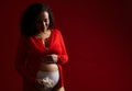 Delightul gorgeous adult pregnant woman smiling, stroking her belly, posing with a white orchid flower, red background