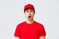 Delivery, contactless orders and shopping concept. Embarrassed and shocked asian courier in red cap and t-shirt uniform
