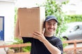 A delivery man with a prominent mustache and a blue hat delivering a generic brown box at a house