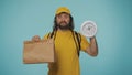 Delivery man in yellow uniform holding a paper bag and clock. Isolated on blue background. Royalty Free Stock Photo