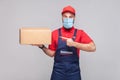 Delivery on quarantine. Young man with surgical medical mask in blue uniform and red t-shirt standing, holding and pointing finger