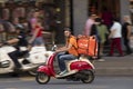 Delivery service courier riding a red and beige vintage vespa scooter in the city street traffic