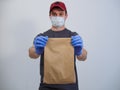 A deliveryman in a red cap in a mask and gloves holds a package with groceries on a white background. Food delivery in