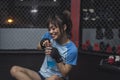 After a demanding training session, a smiling and happy female MMA novice sits down on the ring cage floor and hydrates herself