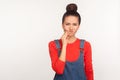 Dental health problems. Portrait of unhealthy girl with hair bun in denim overalls touching sore cheek, suffering toothache