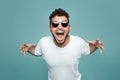 Depressed angry hipster bearded man with sunglasses yelling and screaming, isolated over blue studiou background Royalty Free Stock Photo