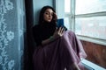 A depressed girl sits at the window with a phone in her hands waiting for a call