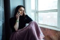 A depressed girl sits at the window with a phone in her hands waiting for a call