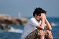 Depressed upset young Asian man covering face with hands sitting on the rock of sea shore Royalty Free Stock Photo