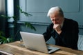 Depressed worried bearded senior aged man sitting at wooden table with laptop compare in dark living room. Royalty Free Stock Photo