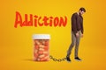 Depressed young man standing and looking down, chained to huge plastic jar of pills on amber background with big red