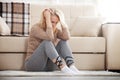 Middle aged barefoot woman sitting at the floor embracing her knees, near sofa at home, her head down, bored, troubled Royalty Free Stock Photo